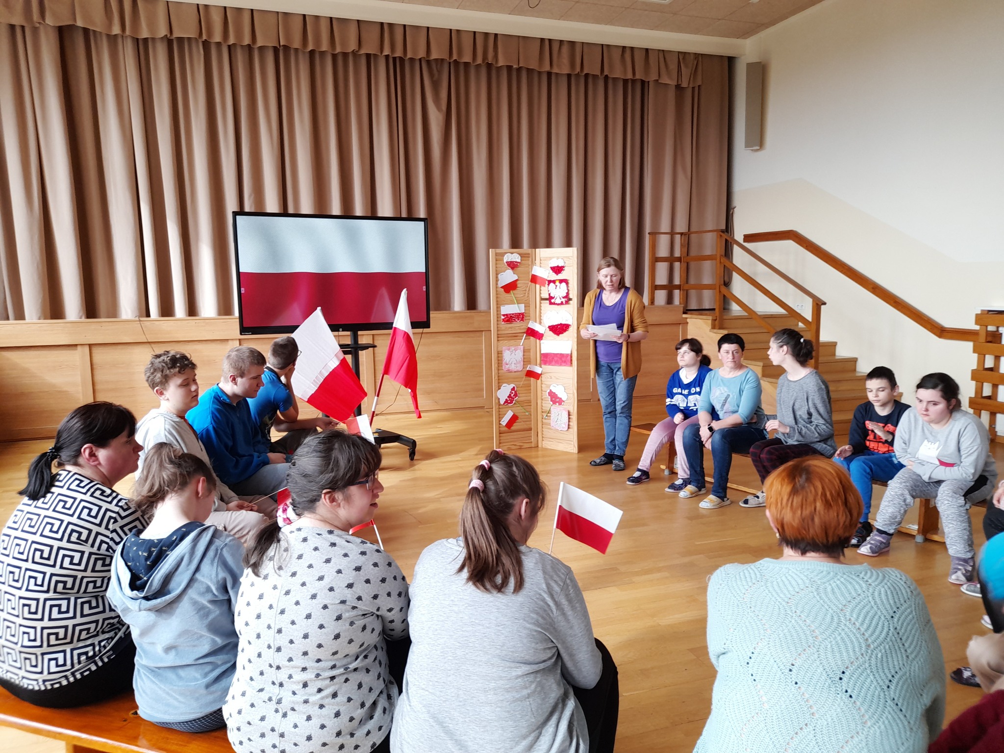 Uczniowie siedzą z flagami polski na ławeczkach i słuchają wychowawcy.