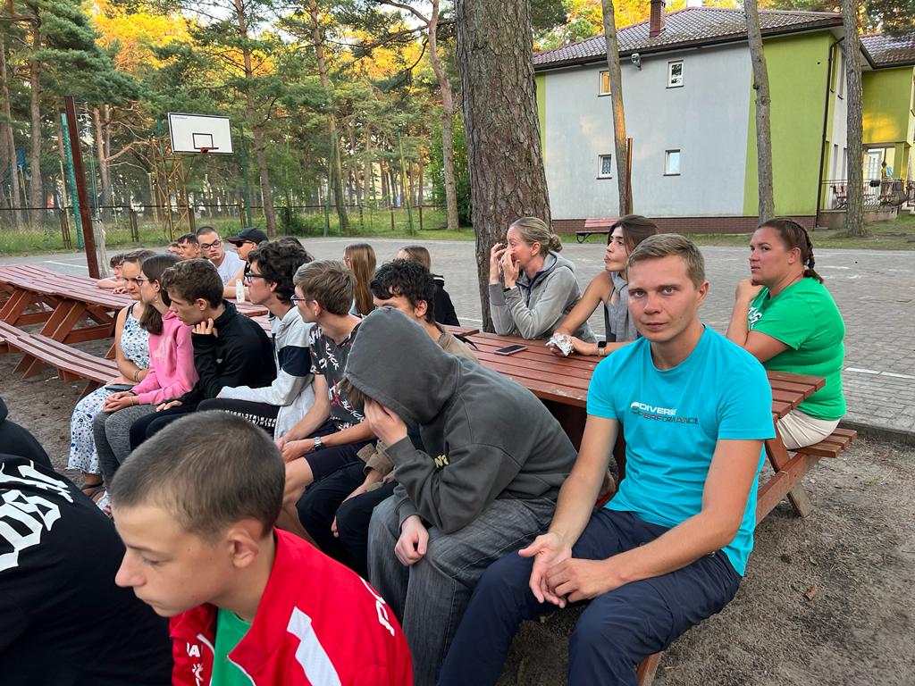 Uczniowie z wychowawcami siedzą na ławkach.