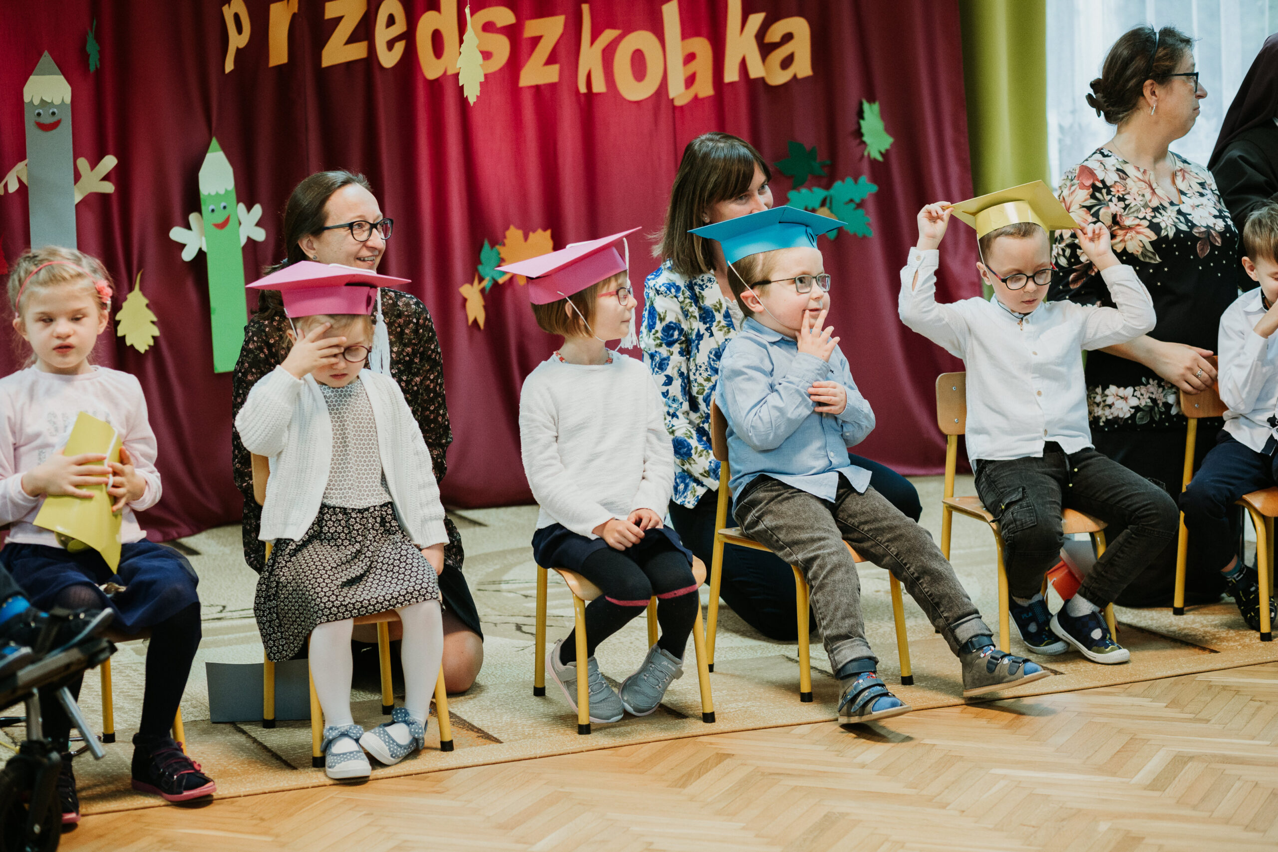 Przedszkolaki w czapkach siedzą na krzesełkach.