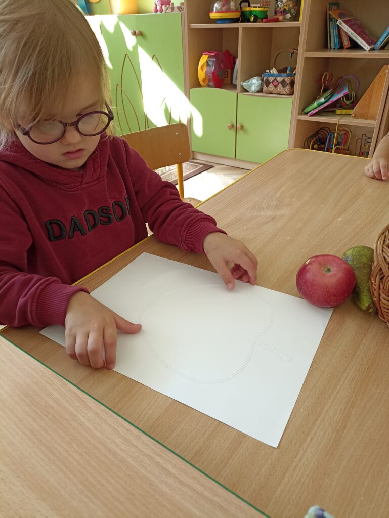 Dziewczynka trzyma kartkę na której są wypukłe kontury w kształcie jabłka.