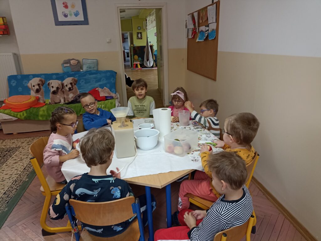 Dzieci siedzą przy stole na którym stoi sokowirówka i miski z owocami.