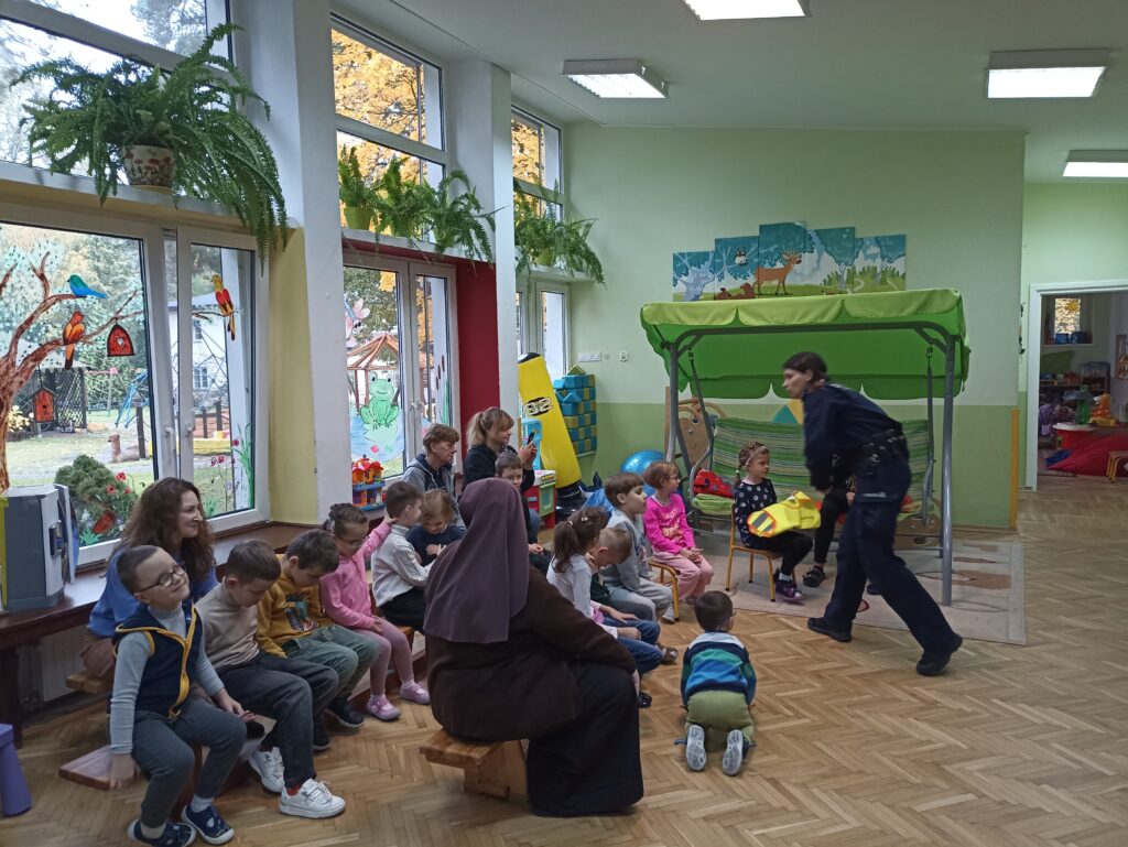 Pani policjantka rozdaje kamizelki odblaskowe dzieci siedzą na ławkach w bawialni w przedszkolu.