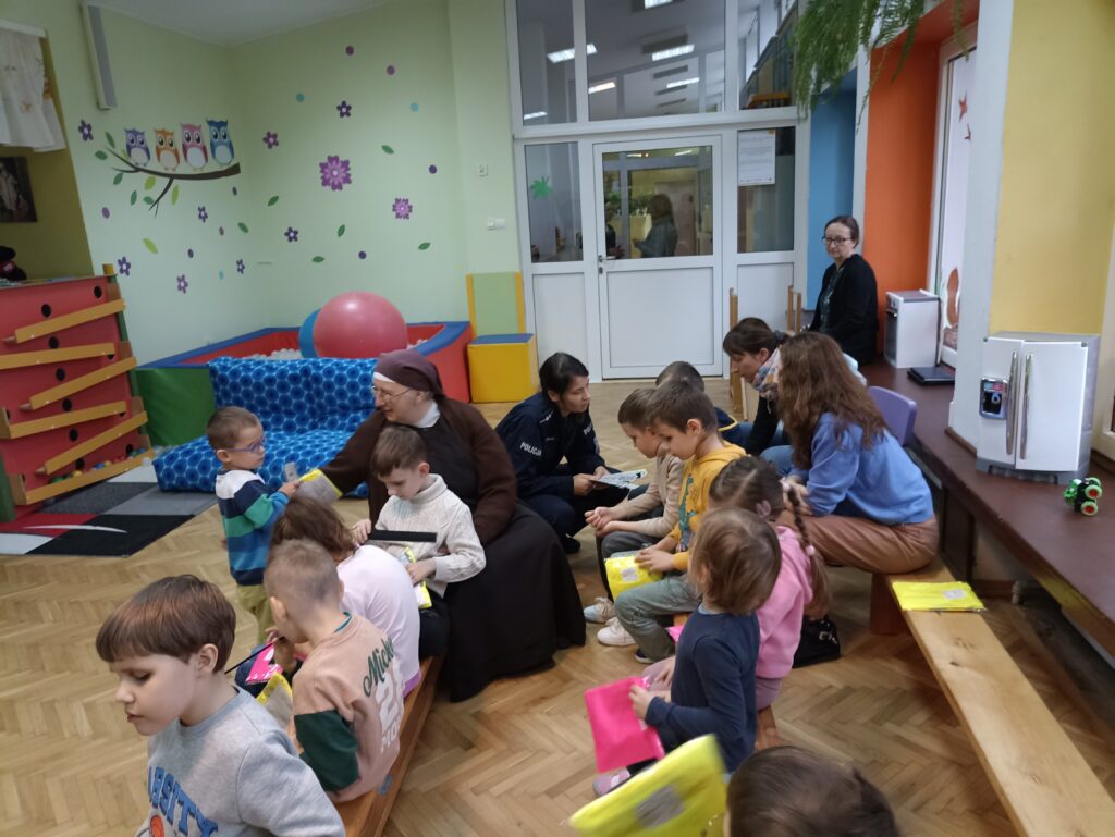 Dzieci z przedszkola i szkoły siedzą na ławkach. Pani policjantka rozdaje kamizelki odblaskowe.