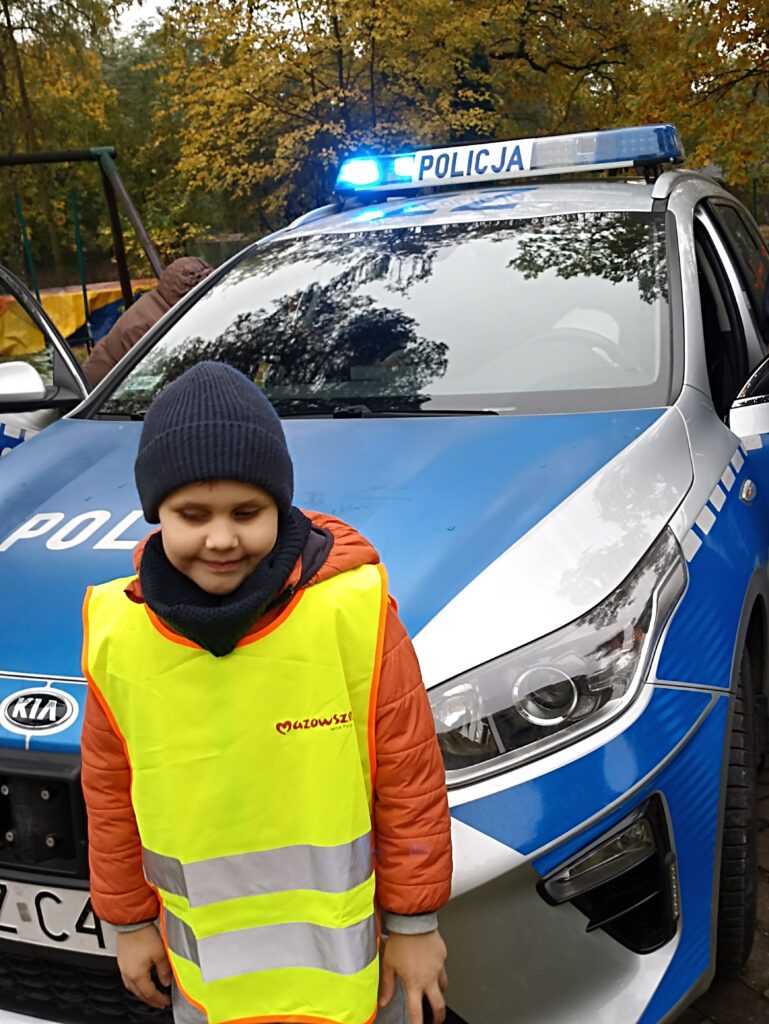 Chłopiec w kamizelce odblaskowej na tle samochodu policyjnego.