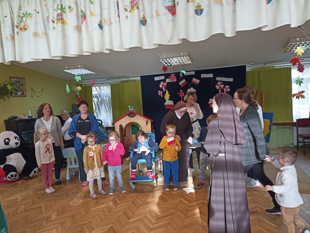 Dzieci i nauczyciele na tle dekoracji patriotycznej - flag i kotylionów w barwach narodowych na ciemnym materiale.