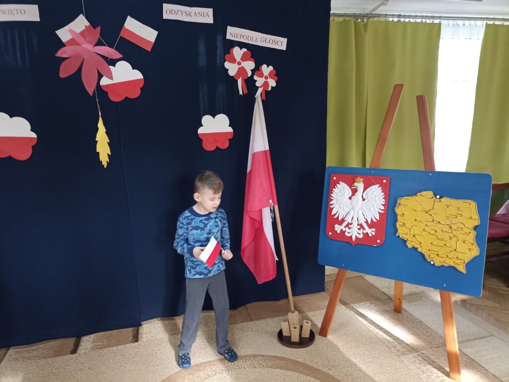 Chłopiec z flagą biało - czerwoną w dłoni na tle dekoracji patriotycznej.