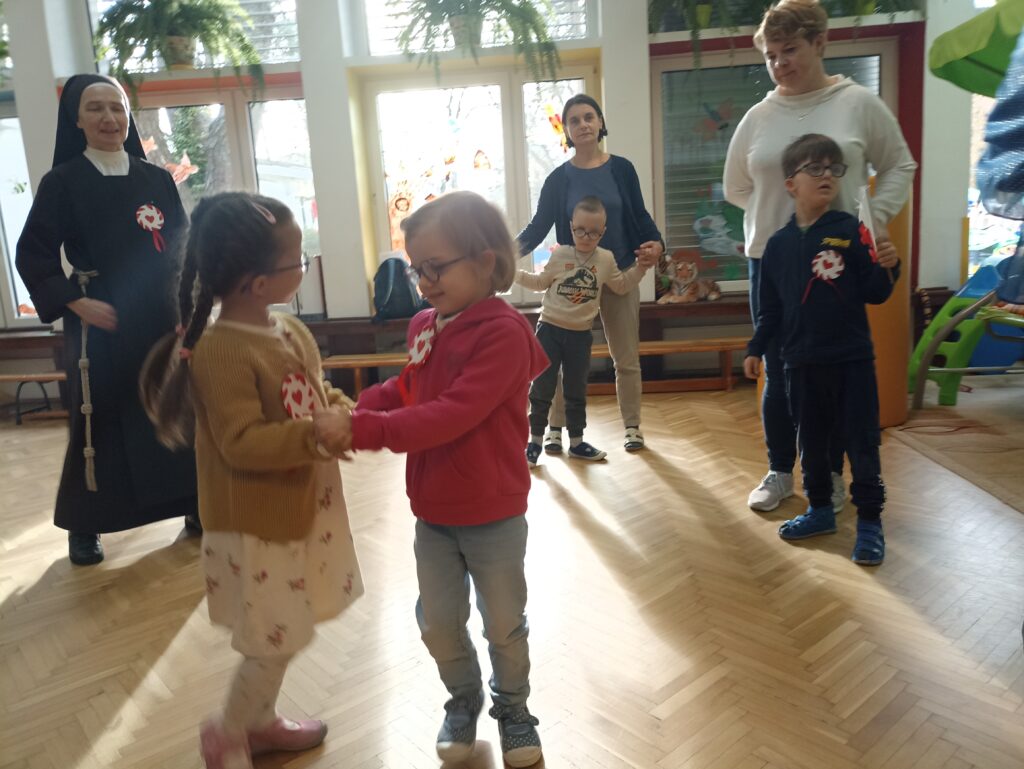 Dwie dziewczynki trzymające się za ręce. W oddali siostra z kotylionem, chłopcy z kotylionami z paniami.