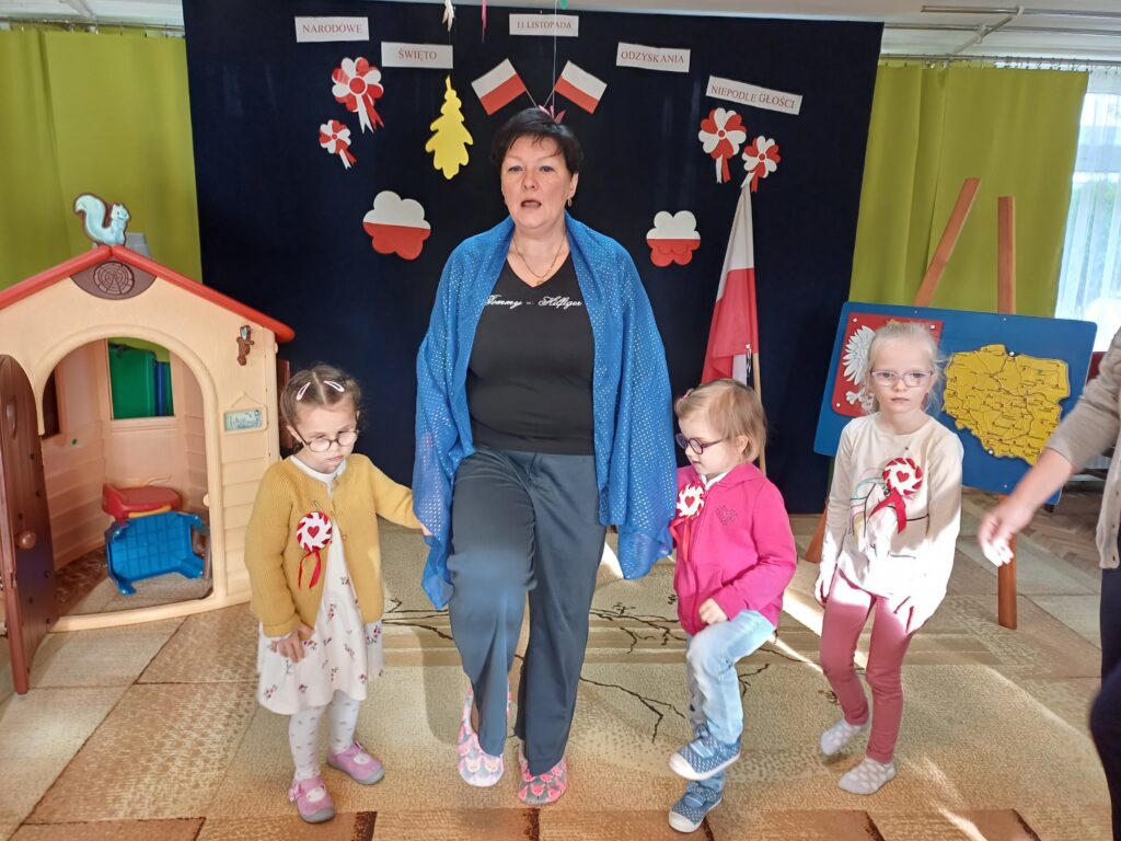 Pani psycholog z trzema dziewczynkami na tle dekoracji patriotycznej. Podnoszą kolana do góry - maszerują. Dziewczynki mają przypięte kotyliony.