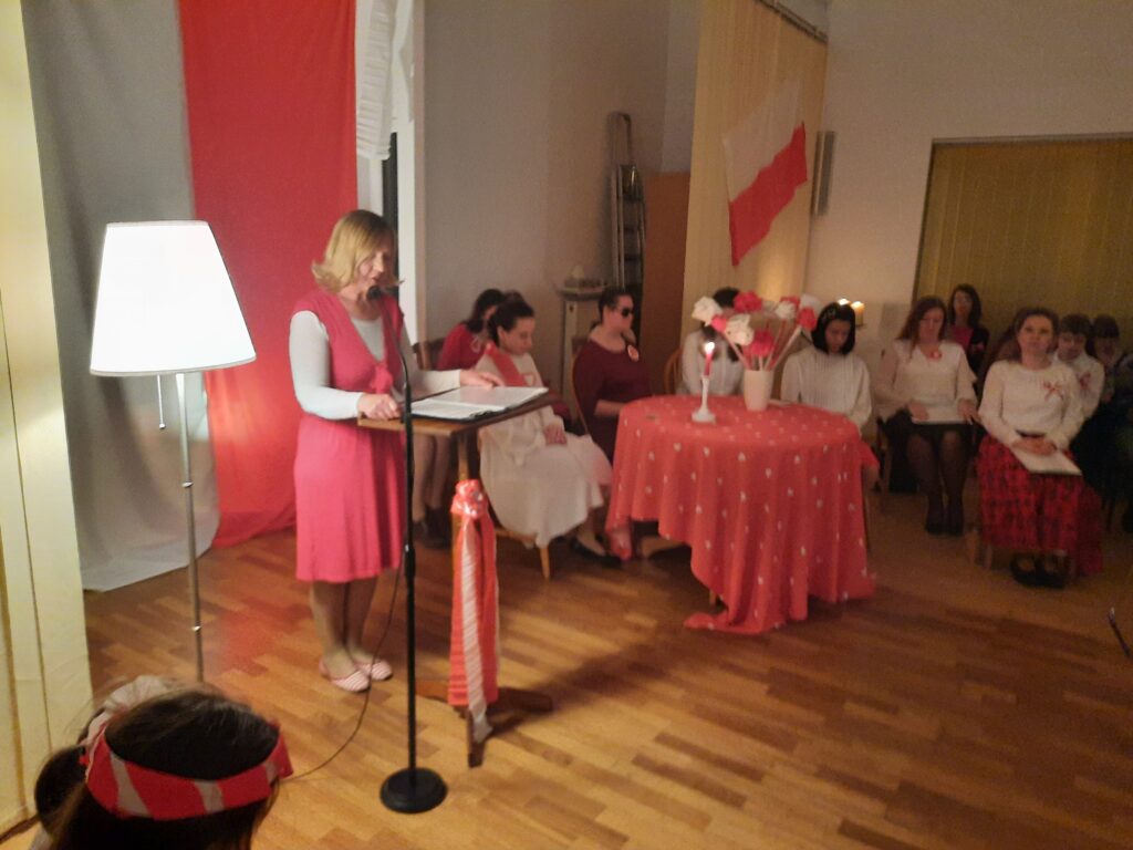 Pani Jola  ubrana w strój biało - czerwony przy mównicy wprowadza  uczestników w podniosłą atmosferę