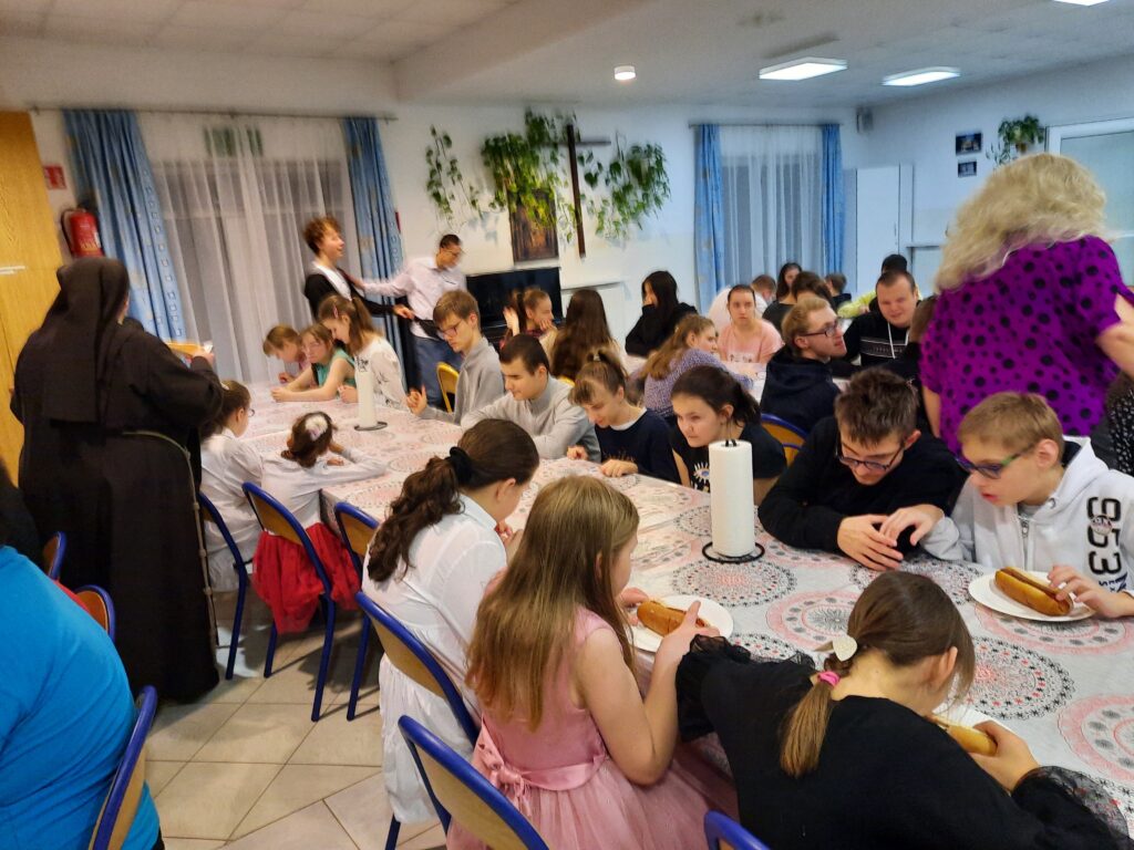 Uczestnicy dyskoteki siedzą przy stołach i jedzą hot dogi