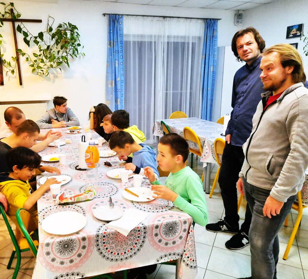 Chłopcy siedzą przy stole i jedzą kolację a nad nimi czuwają wychowawcy