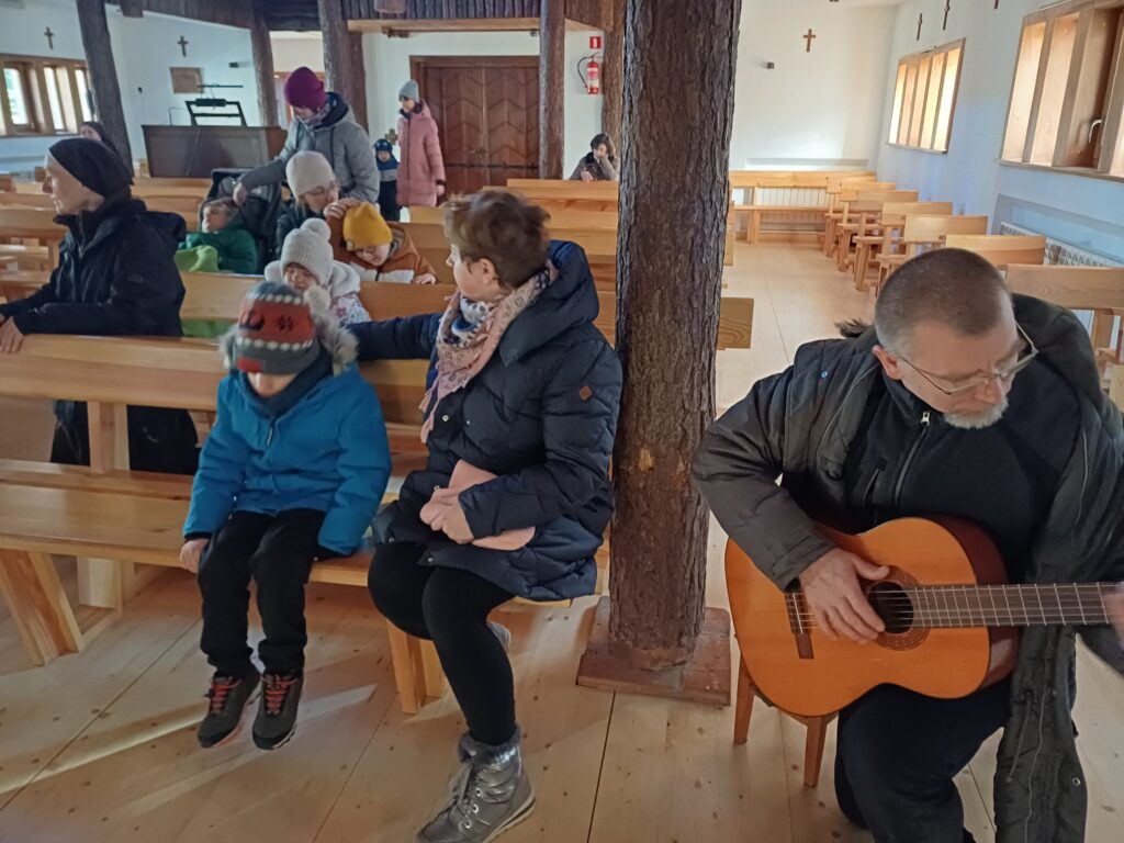 Grupa Krasnali i siostra kierowniczka siedzą w ławkach kościelnych. Pan z gitarą siedzi obok ławek.