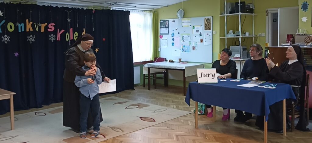 Chłopiec wspierany przez Siostrę śpiewa kolędę. Jury słucha.