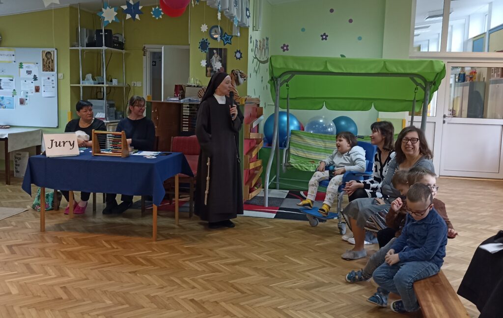 Siostra Dyrektor na stojąco ogłasza wyniki konkursu.  Jury i publiczność słucha. 