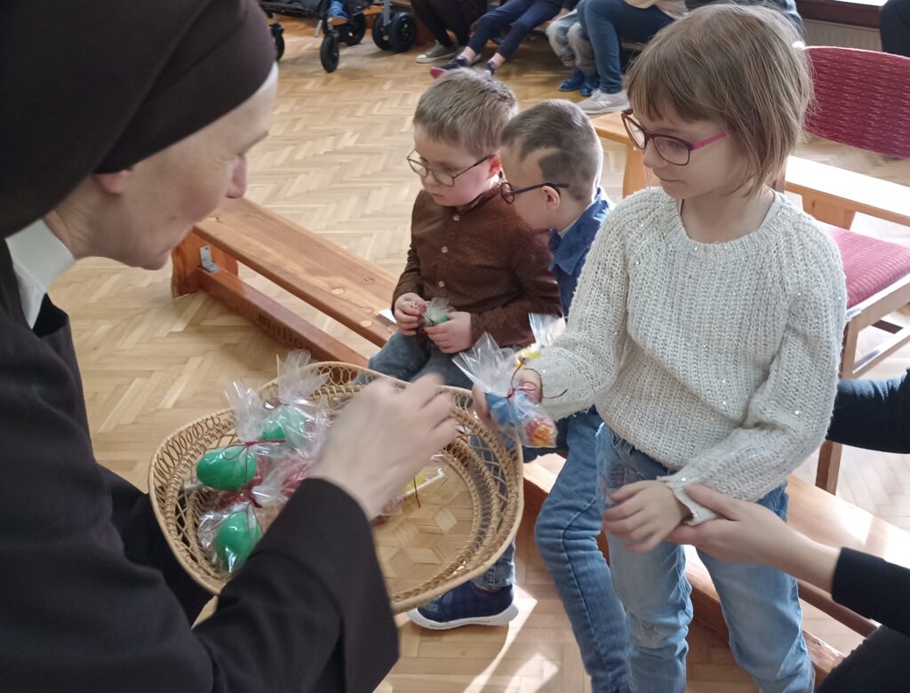 Siostra wręcza dziewczynce nagrodę. Obok siedzi na ławeczce dwóch chłopców z nagrodami w dłoniach.