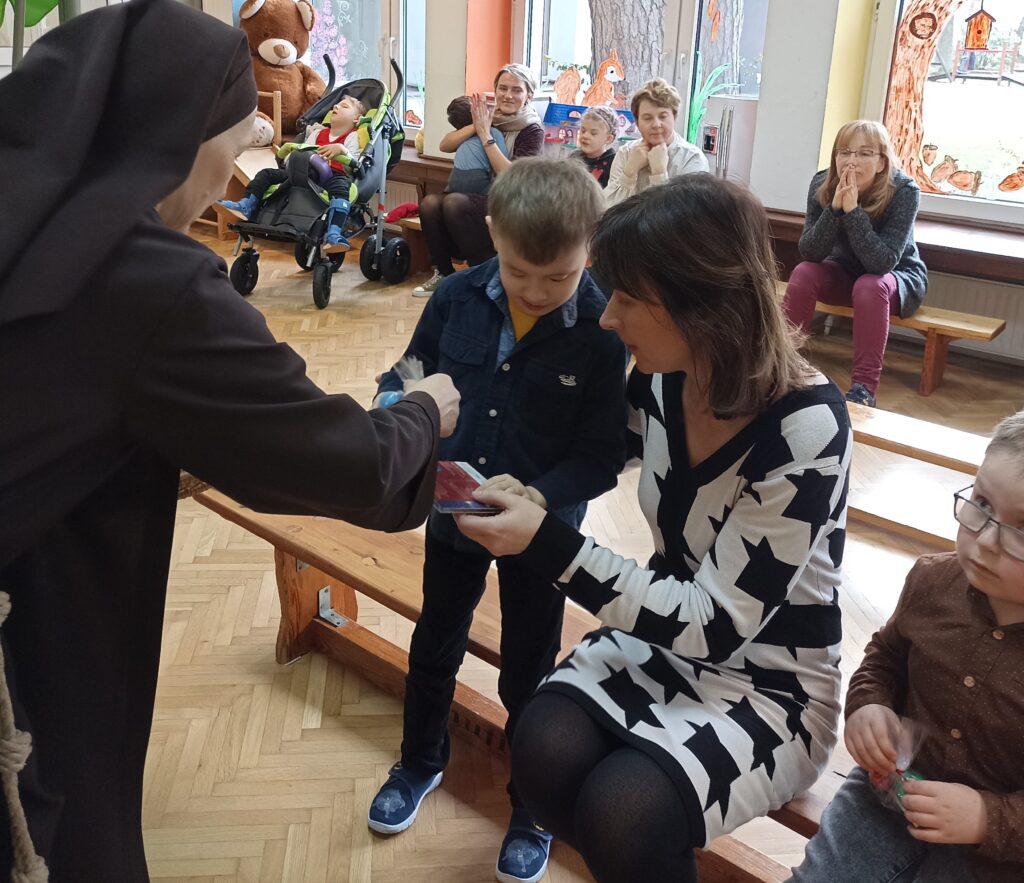 Siostra wręcza na ręce chłopca nagrodę dla grupy Zuchów. Obok nauczycielka prowadząca grupę pomaga chłopcu odebrać nagrodę. W tle inne dzieci i nauczycielki. 
