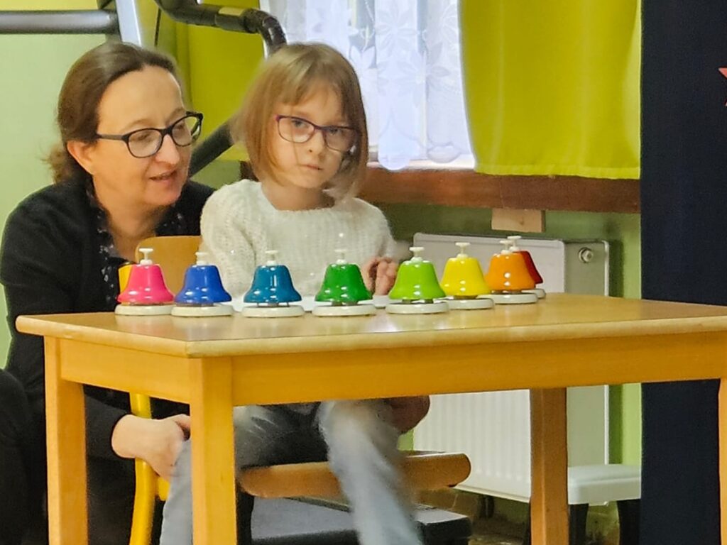 Dziewczynka siedzi przy stoliku, na którym ustawione są dzwonki naciskane. Obok siedzi nauczycielka.