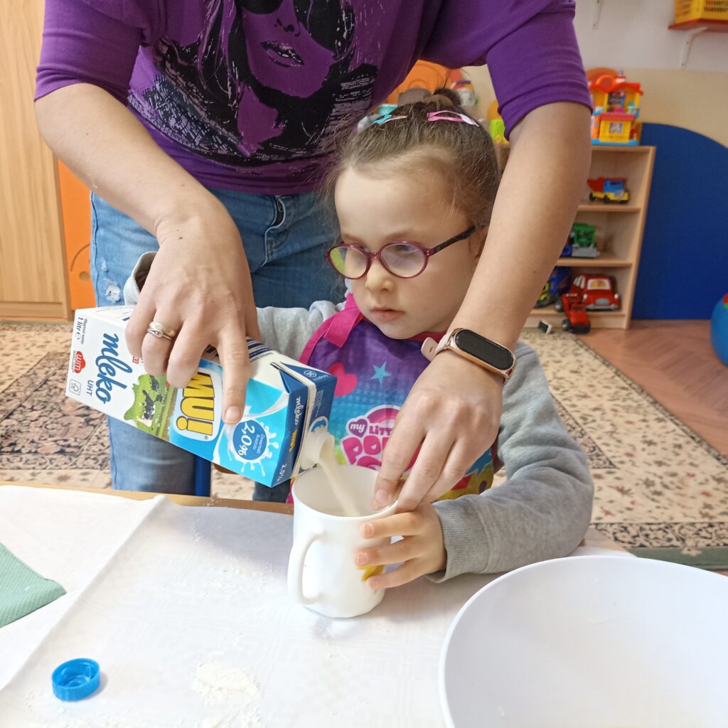 Dziewczynka wlewa mleko z pomocą Pani.
