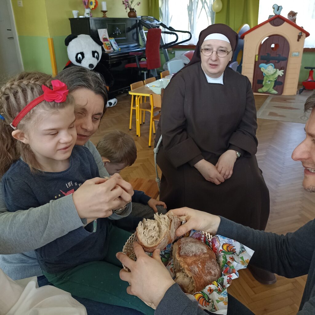 Ksiądz podaje dziewczynce chleb, w tle s. Adriana.