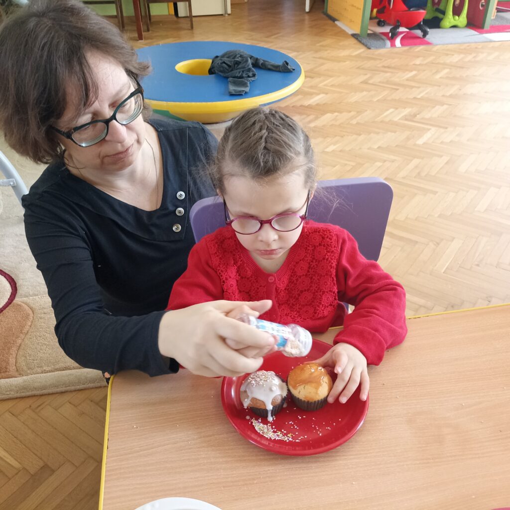 Dziewczynka z pomocą Pani dekoruje babeczkę.