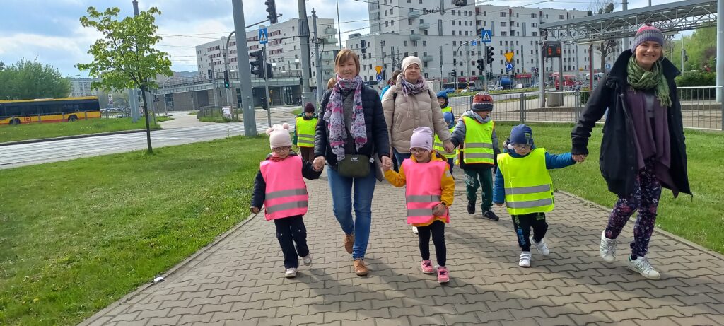 Przedszkolaki za rękę z opiekunami idą chodnikiem ubrane w kamizelki odblaskowe.