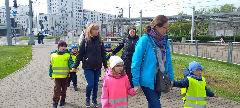 Przedszkolaki za rękę z opiekunami idą chodnikiem ubrane w kamizelki odblaskowe.