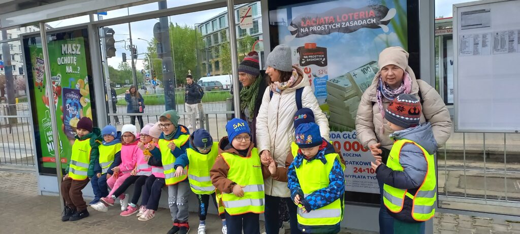 Przedszkolaki w kamizelkach odblaskowych z opiekunami na przystanku tramwajowym.