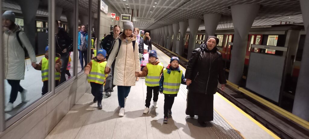 Przedszkolaki w kamizelkach odblaskowych z opiekunkami idą wzdłuż peronu metra.