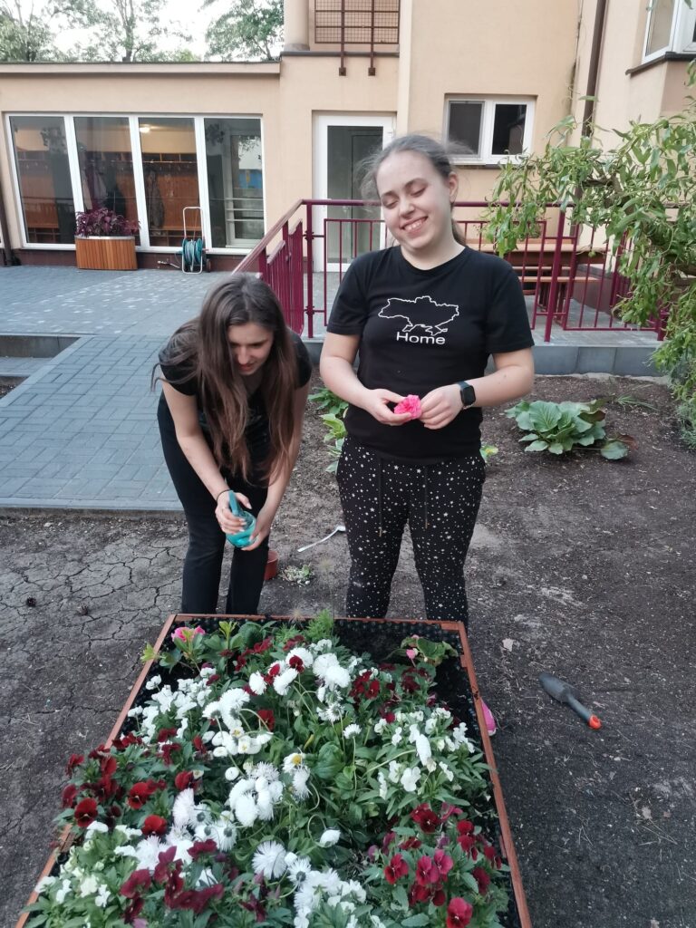 Nikola podlewa wodą kwiaty a Amina sadzi różową begonię