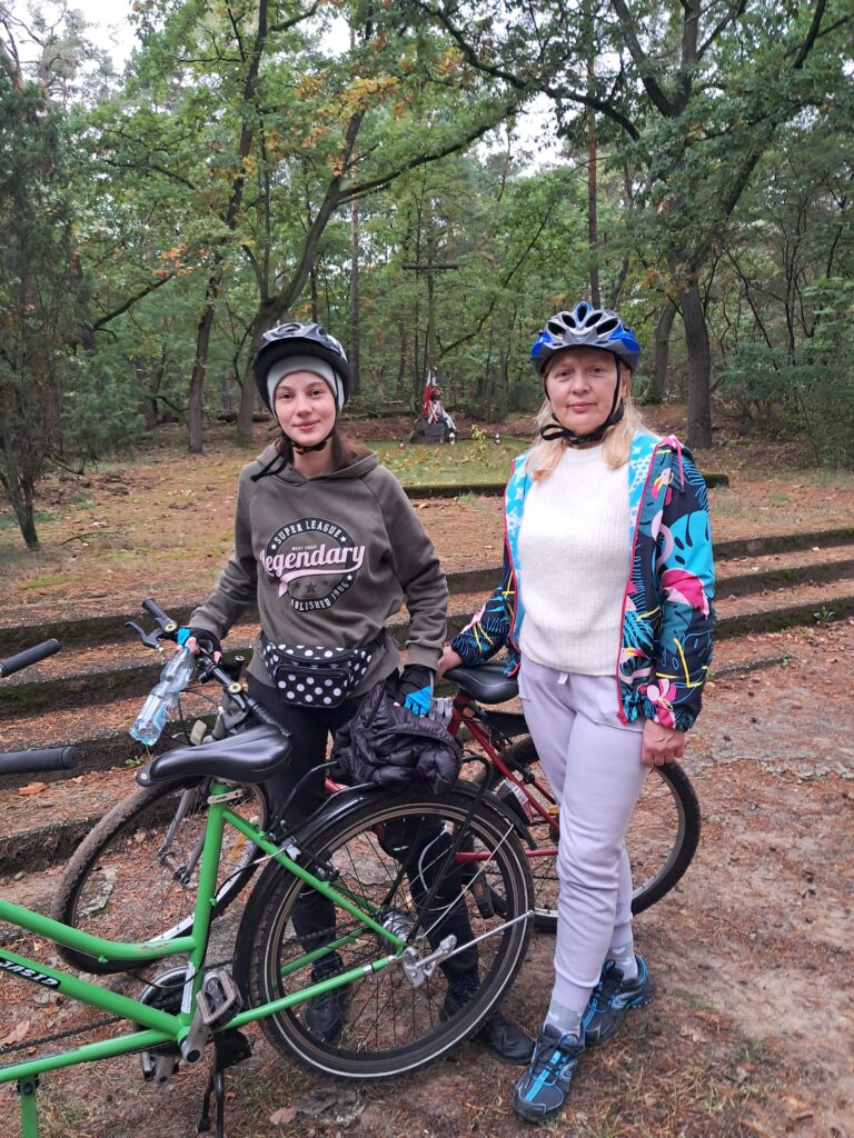 Pani Jola z Valerią w strojach sportowych stoją przy rowerach a oddali widać Mogiłę Powstańców