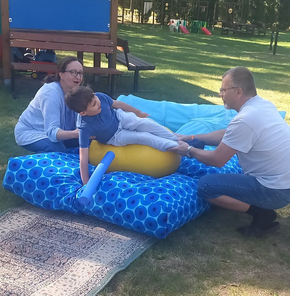 Rehabilitant i nauczycielka asekurują chłopca podczas zabawy na ruchomej piłce fasolce symulującej statek na morzu.