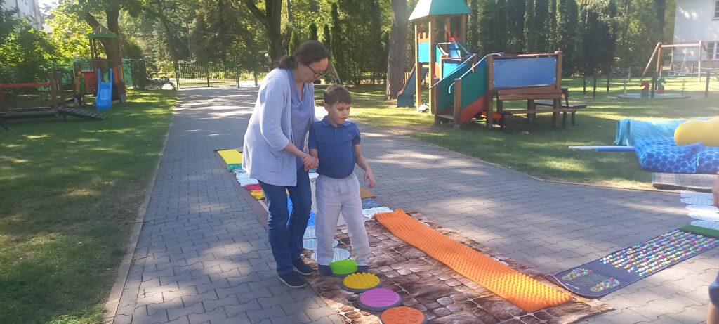 Nauczycielka pomaga chłopcu przejść po ścieżce sensorycznej
