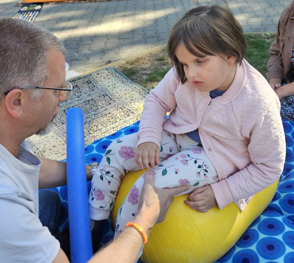 Rehabilitant asekuruje dziewczynkę podczas zabawy na ruchomej piłce fasolce symulującej statek na morzu.