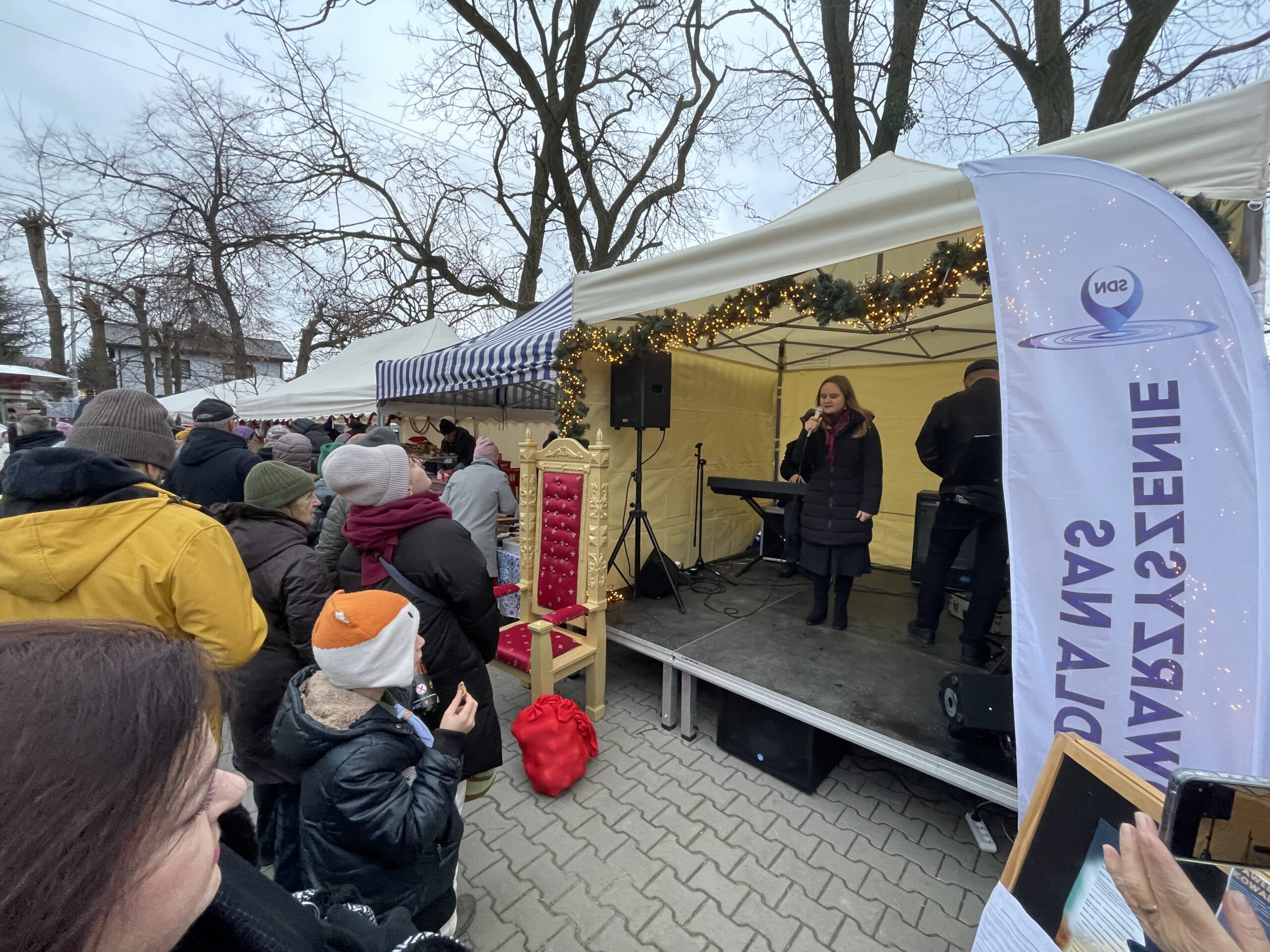 Julia śpiewa na scenie, przed którą stoją osoby odwiedzające ryneczek.