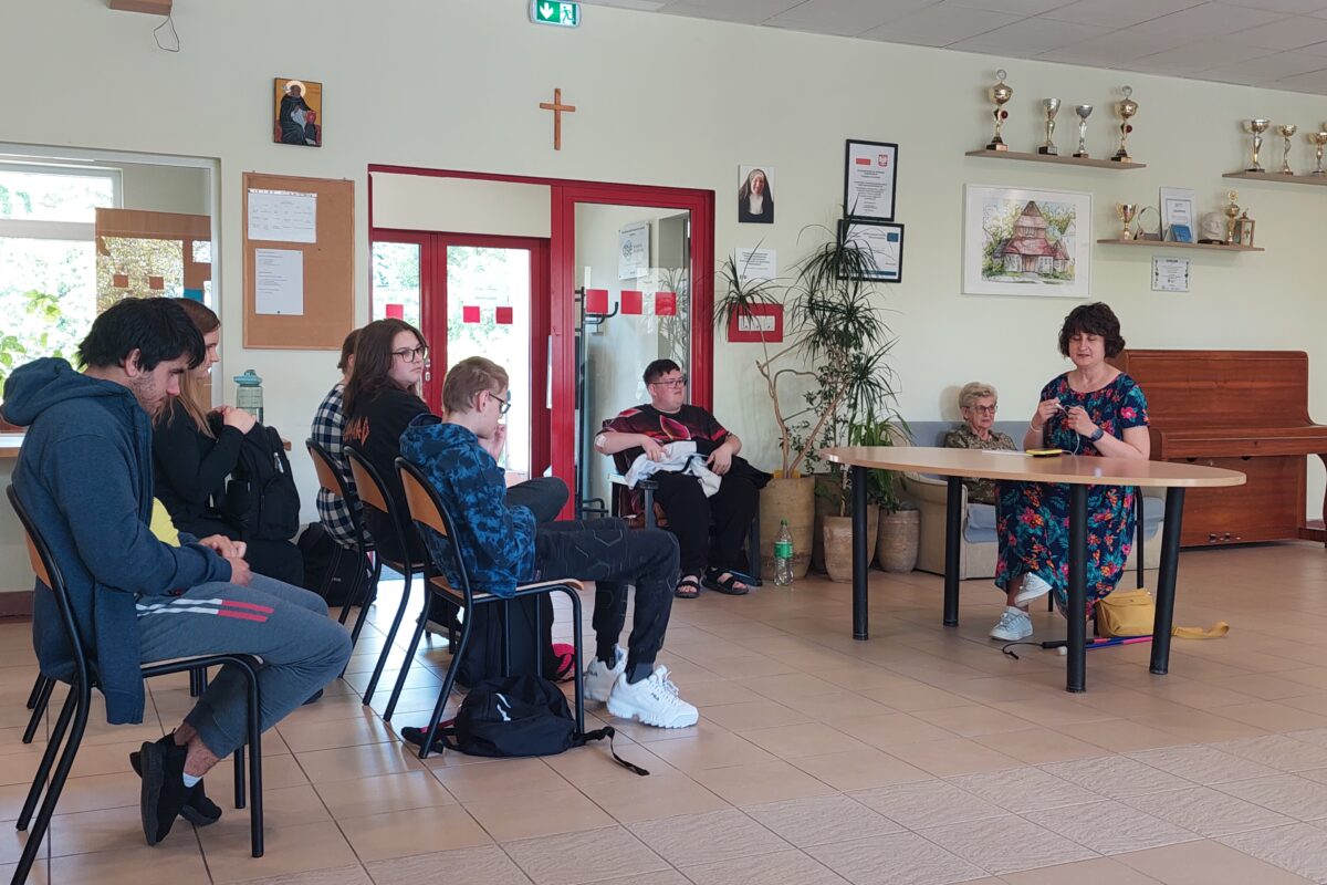Małgorzata siedzi przy stoliku skierowana do siedzących przed nią w rzędach uczniów i nauczycieli w Szkole Ponadpodstawowej