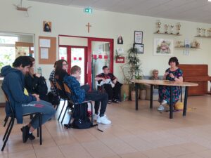 Małgorzata siedzi przy stoliku skierowana do siedzących przed nią w rzędach uczniów i nauczycieli w Szkole Ponadpodstawowej
